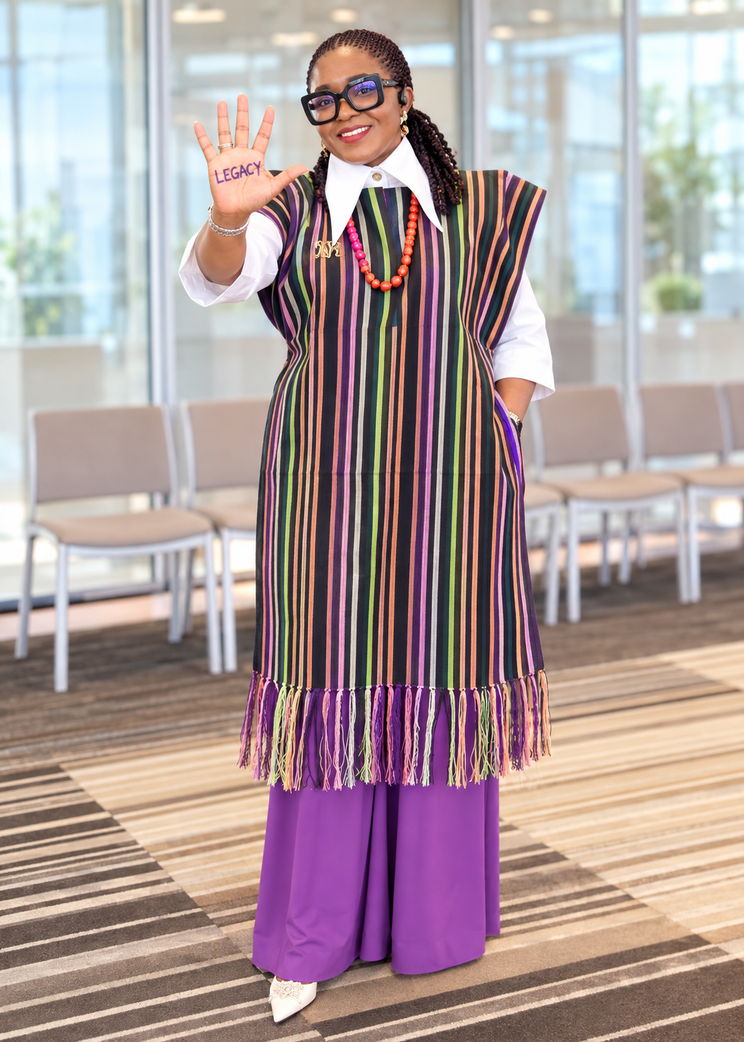 Adedamola in a striped woven outfit and purple trousers, raising her hand with the word Legacy.